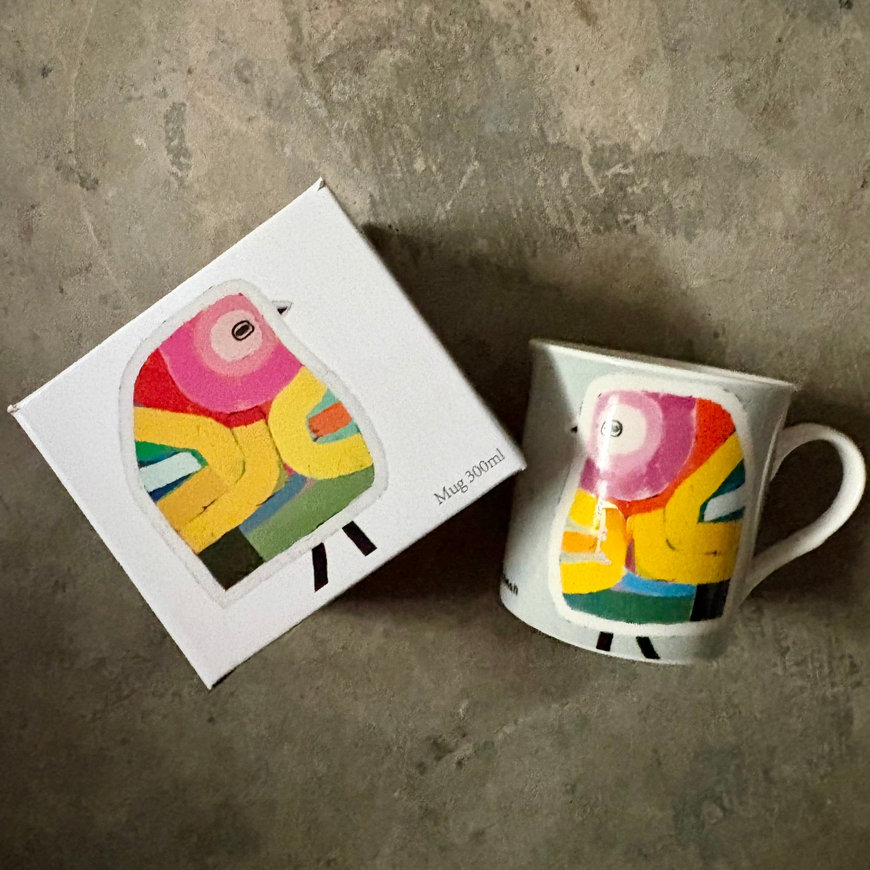 Lilli Rock Dolly Mug - Colourful mug with a bird design next to its gift box on a concrete surface. 