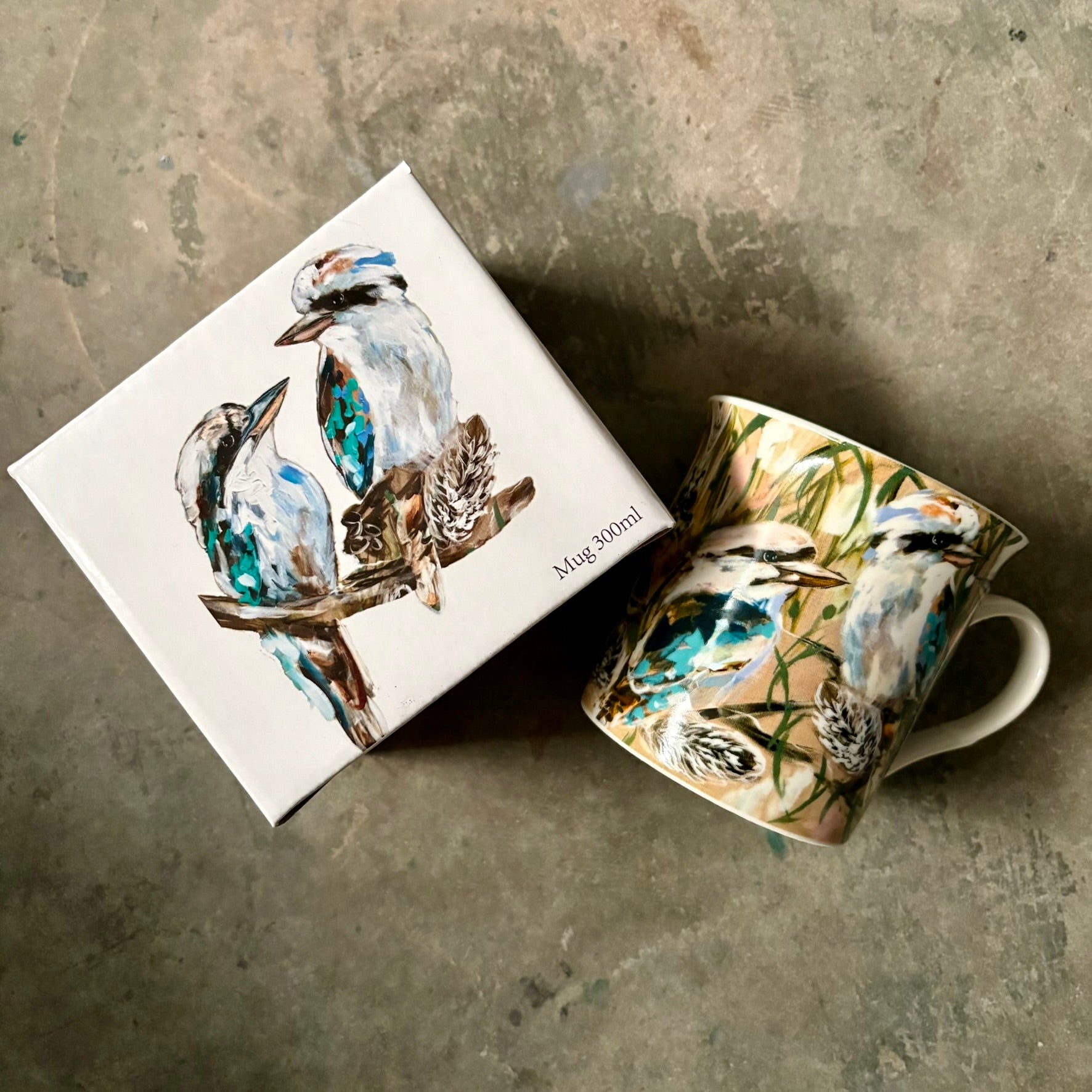 Lilli Rock Kookaburra & Banksia Mug - A ceramic mug and its matching square gift box, both decorated with a watercolour-style painting of kookaburras perched on branches. 